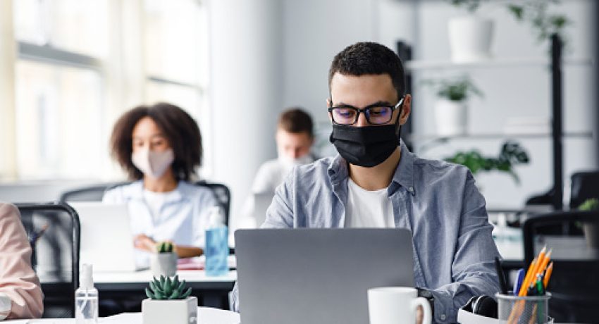 Office center workers are protected from virus outbreak during covid-19 epidemic. Young hipster man in glasses and protective mask works at laptop, with colleagues at workplaces in interior, free space