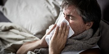 Sick man with flu lying in bed and blow nose napkin.