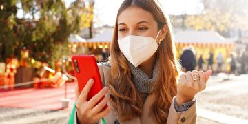 Excited young woman wearing medical mask FFP2 KN95 watching smartphone and holding shopping bags walking in the Christmas Markets