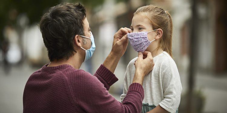 Father with surgical face mask, is putting a hand made protective face mask on his little daughter for the COVID-19 pandemic.